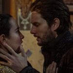 Cena intensa mostrando os personagens Darkling e Alina frente a frente em um momento de tensão e emoção, prestes a se beijar. Ele segura o rosto dela com as duas mãos enquanto ela o encara, em um ambiente com iluminação suave e detalhes dourados ao fundo.