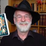 O autor da Terry Pratchett aparece sorrindo, usando chapéu preto, óculos e barba branca, posando em uma biblioteca com estantes cheias de livros ao fundo. Ao lado dele está a capa do livro “Os Pequenos Homens Livres”, ilustrada com elementos de fantasia como cogumelos, criaturas pequenas e uma jovem segurando uma lupa.